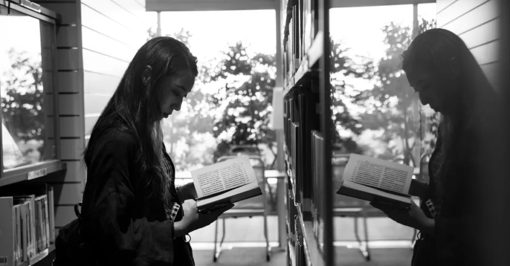 Seorang wanita yang khusyuk membaca buku di samping jendela reflektif di perpustakaan.