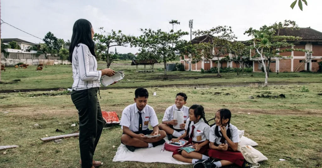 Siswa berpakaian seragam menikmati kelas luar ruangan bersama guru di taman.