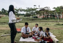 Siswa berpakaian seragam menikmati kelas luar ruangan bersama guru di taman.