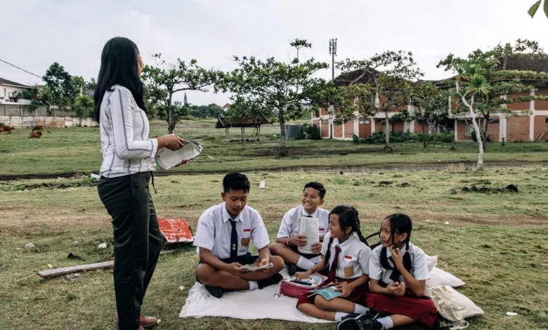 Siswa berpakaian seragam menikmati kelas luar ruangan bersama guru di taman.