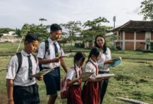 Sekelompok siswa mengenakan seragam sekolah membaca di luar ruangan bersama guru mereka di hari yang cerah.