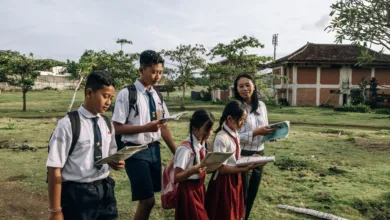 Sekelompok siswa mengenakan seragam sekolah membaca di luar ruangan bersama guru mereka di hari yang cerah.