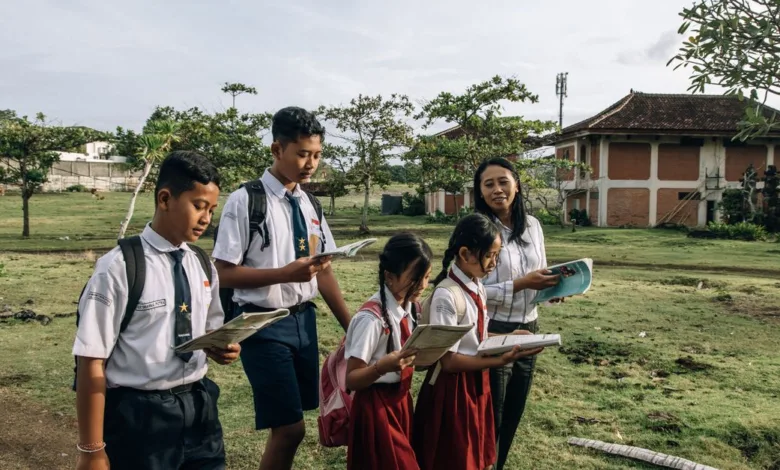 Sekelompok siswa mengenakan seragam sekolah membaca di luar ruangan bersama guru mereka di hari yang cerah.