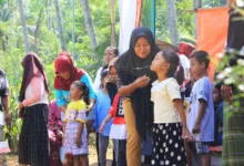 Pertemuan luar ruangan yang meriah dengan anak-anak dan orang dewasa berinteraksi di bawah sinar matahari dekat pohon.