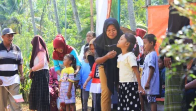 Pertemuan luar ruangan yang meriah dengan anak-anak dan orang dewasa berinteraksi di bawah sinar matahari dekat pohon.