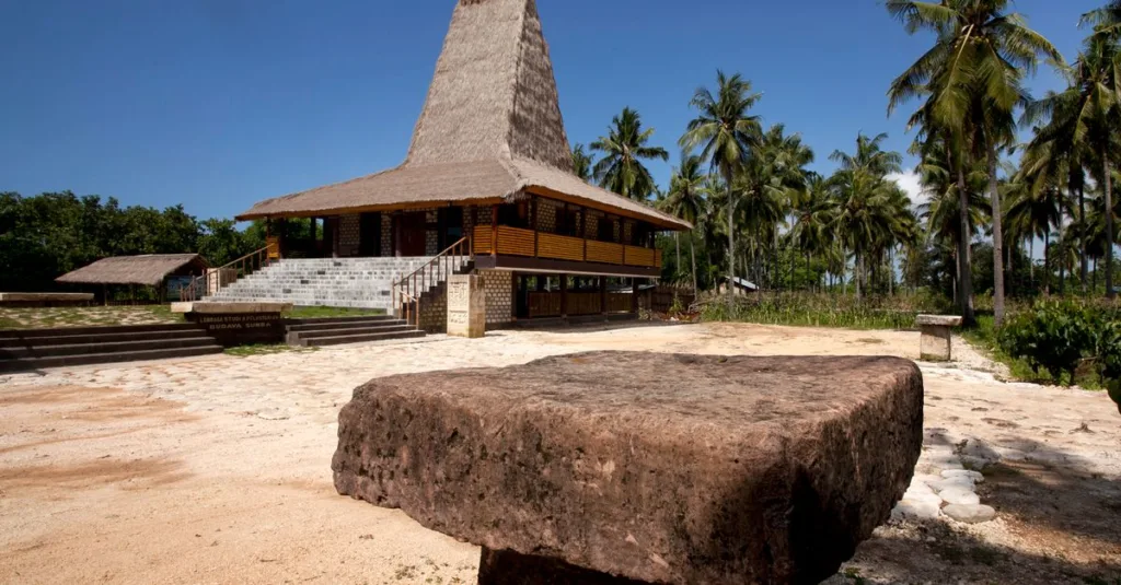 Jelajahi rumah tradisional Sumbawa yang dikelilingi pohon palem rimbun di bawah langit biru cerah.