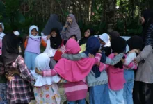 Sekelompok anak-anak yang gembira berpartisipasi dalam kegiatan luar ruangan yang dipandu oleh orang dewasa di lingkungan hijau yang rimbun.
