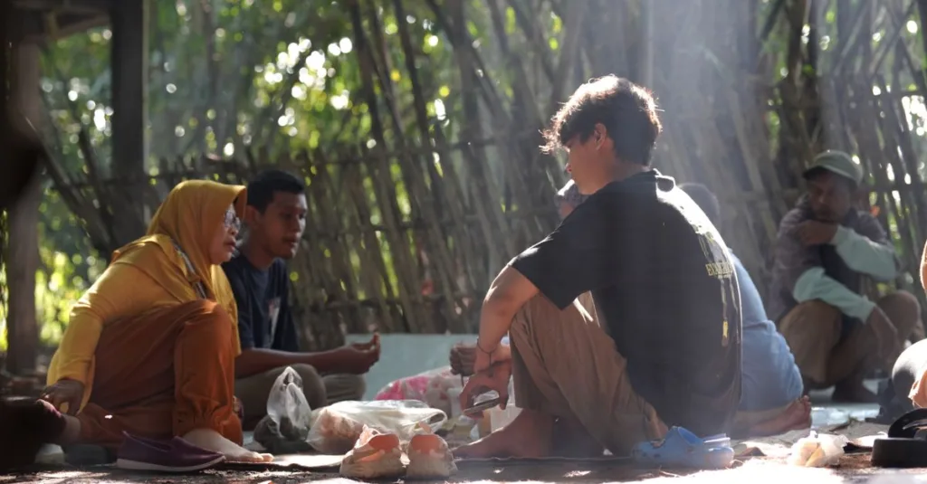 Sekelompok orang berkumpul di luar ruangan di alam, terlibat dalam percakapan dan berbagi makanan.