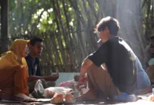 Sekelompok orang berkumpul di luar ruangan di alam, terlibat dalam percakapan dan berbagi makanan.