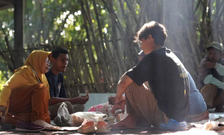 Sekelompok orang berkumpul di luar ruangan di alam, terlibat dalam percakapan dan berbagi makanan.