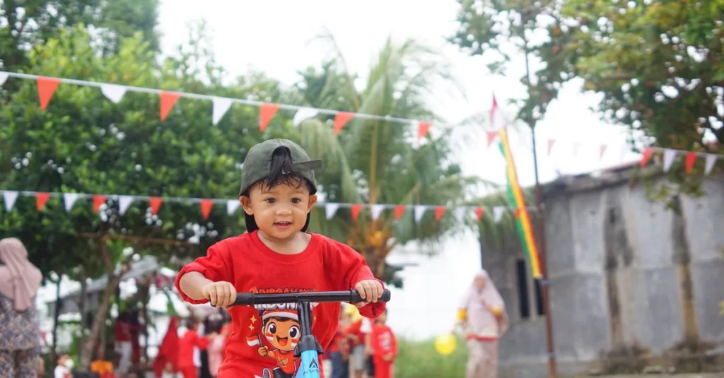 Seorang anak mengendarai skuter saat festival luar ruangan di Indonesia, dengan dekorasi meriah dan orang-orang di latar belakang.