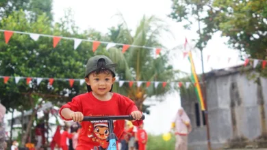 Seorang anak mengendarai skuter saat festival luar ruangan di Indonesia, dengan dekorasi meriah dan orang-orang di latar belakang.