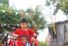Seorang anak mengendarai skuter saat festival luar ruangan di Indonesia, dengan dekorasi meriah dan orang-orang di latar belakang.