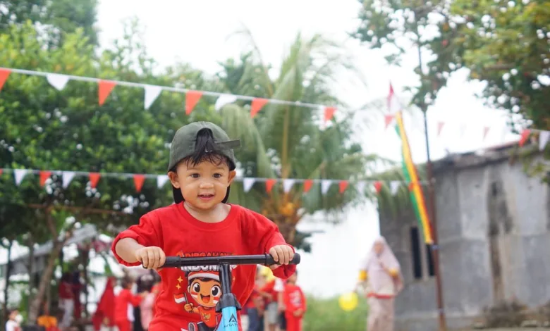 Seorang anak mengendarai skuter saat festival luar ruangan di Indonesia, dengan dekorasi meriah dan orang-orang di latar belakang.