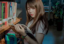 Seorang wanita muda membaca dengan penuh perhatian di lorong perpustakaan, dikelilingi oleh buku-buku.