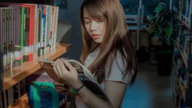 Seorang wanita muda membaca dengan penuh perhatian di lorong perpustakaan, dikelilingi oleh buku-buku.