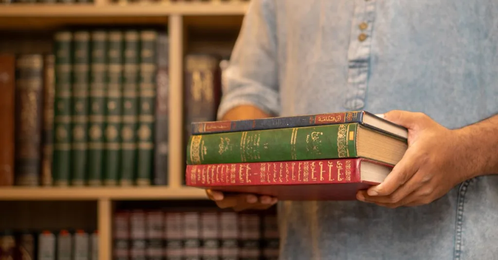 Seorang pria mengenakan kemeja denim memegang tumpukan buku berbahasa Arab di sebuah perpustakaan, melambangkan studi dan pembelajaran.