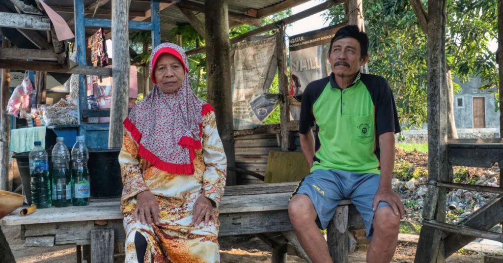 Pasangan lansia duduk di luar ruangan dalam suasana desa Indonesia yang rustic, mencerminkan gaya hidup yang damai.