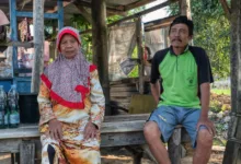 Pasangan lansia duduk di luar ruangan dalam suasana desa Indonesia yang rustic, mencerminkan gaya hidup yang damai.