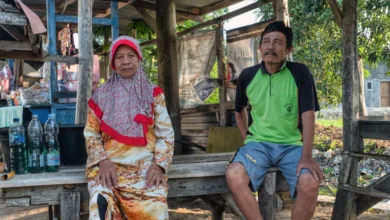 Pasangan lansia duduk di luar ruangan dalam suasana desa Indonesia yang rustic, mencerminkan gaya hidup yang damai.