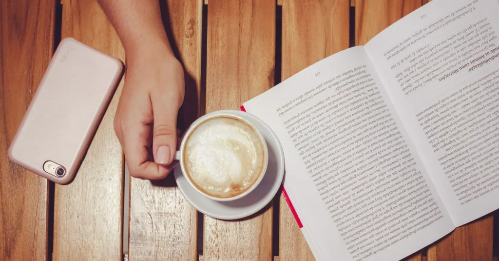 Sebuah pemandangan damai yang menampilkan sebuah buku terbuka, secangkir latte, dan smartphone di atas meja kayu.