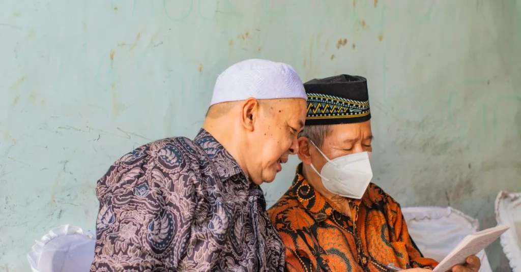 Dua pria paruh baya dalam pakaian tradisional membaca dokumen di dalam ruangan, salah satunya mengenakan masker wajah.