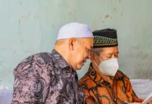Dua pria paruh baya dalam pakaian tradisional membaca dokumen di dalam ruangan, salah satunya mengenakan masker wajah.