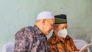 Dua pria paruh baya dalam pakaian tradisional membaca dokumen di dalam ruangan, salah satunya mengenakan masker wajah.
