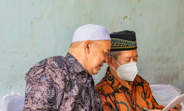 Dua pria paruh baya dalam pakaian tradisional membaca dokumen di dalam ruangan, salah satunya mengenakan masker wajah.